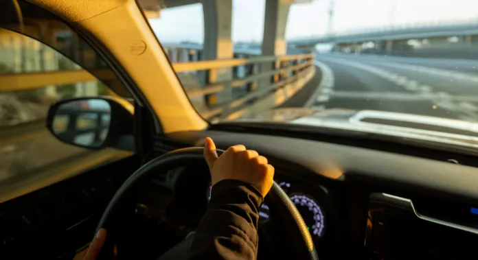 Mão de motorista segurando o volante enquanto dirige em uma rodovia com curva e estruturas elevadas ao fundo, sob a luz do pôr do sol.